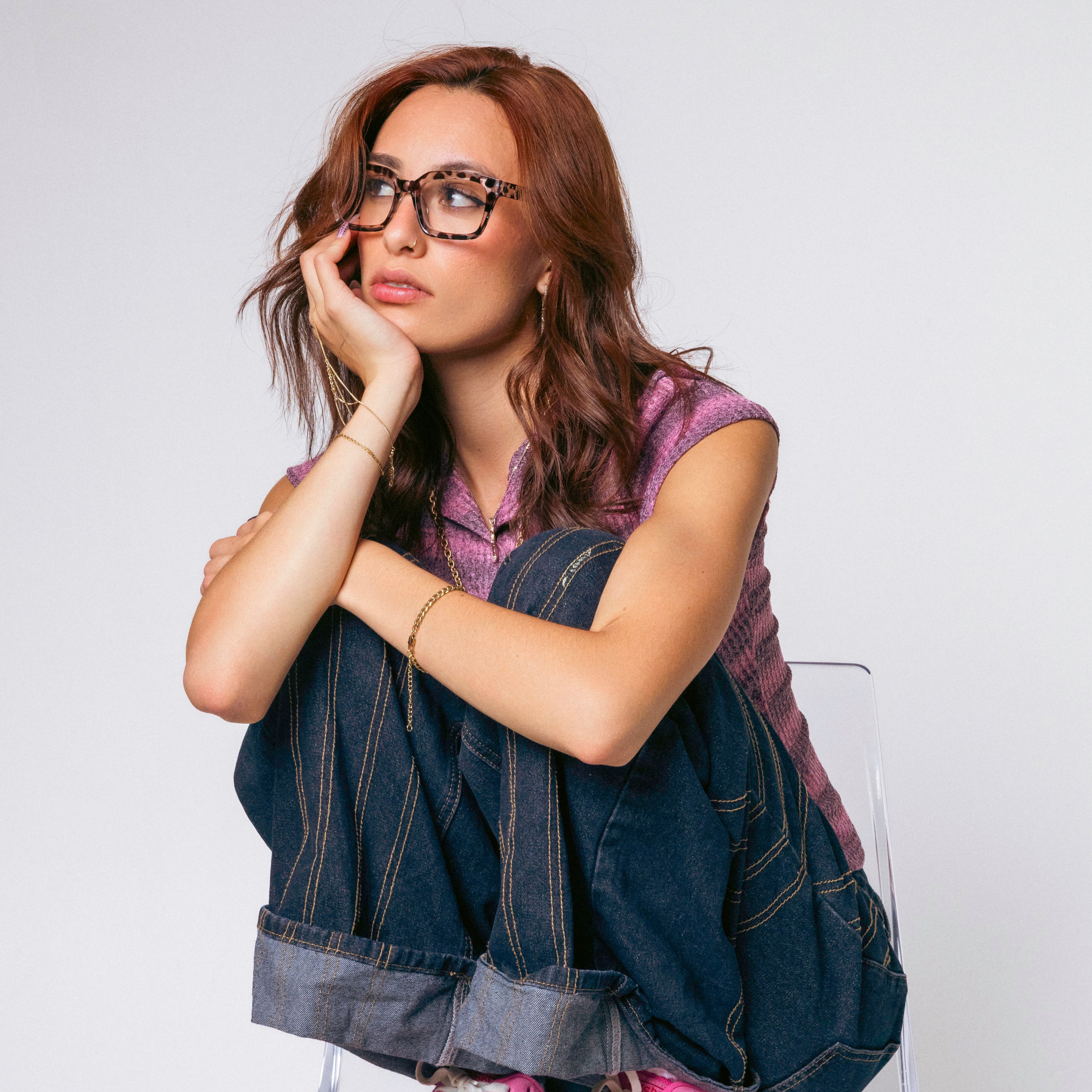 Woman sitting on a clear chair wearing pink sneakers, a denim jacket, and a purple shirt against a white background.